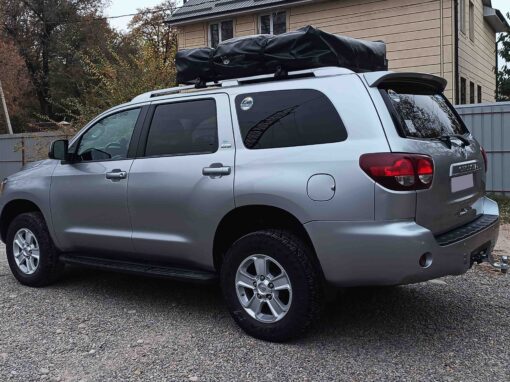 Un SUV Toyota Sequoia argenté, équipé d'un sac de rangement fixé sur le toit, garé sur une allée en gravier dans un décor de 4x4 au Kirghizistan.