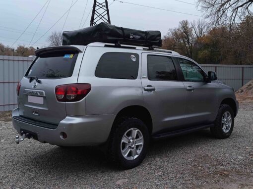 Un SUV Toyota Sequoia argenté, équipé d'une tente de toit noire, garé sur un parking en gravier lors d'un voyage en 4x4 au Kirghizistan.