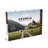 Le Guide de voyage Explore Géorgie présente une image panoramique d'une voiture se dirigeant vers d'anciennes ruines en pierre dans des collines verdoyantes, avec des montagnes majestueuses à l'arrière-plan.