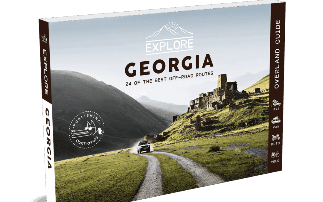 Le Guide de voyage Explore Géorgie présente une image panoramique d'une voiture se dirigeant vers d'anciennes ruines en pierre dans des collines verdoyantes, avec des montagnes majestueuses à l'arrière-plan.