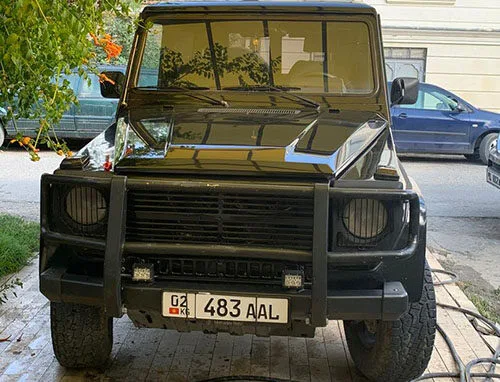 A black off-road vehicle with a front bull bar is parked on a tiled driveway. The license plate reads 483 AAL. There are trees and other parked cars in the background.