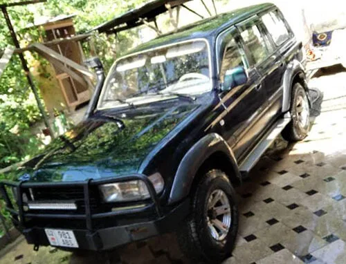 A dark green SUV with a front bull bar is parked on a tiled driveway outdoors, surrounded by greenery and trees in the background.