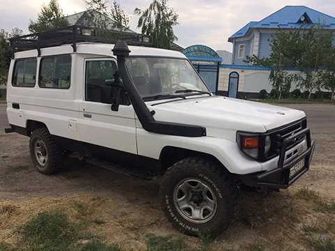 A white off-road SUV with a roof rack and snorkel is parked on a patch of dirt near a paved road, with a blue-roofed house and trees in the background.