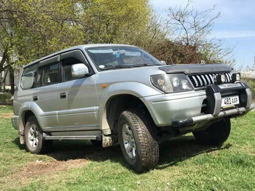 A silver Toyota Land Cruiser Prado SUV is parked on grassy terrain with one front wheel raised on a small mound. Trees with green foliage are in the background, and the vehicle’s license plate is visible.