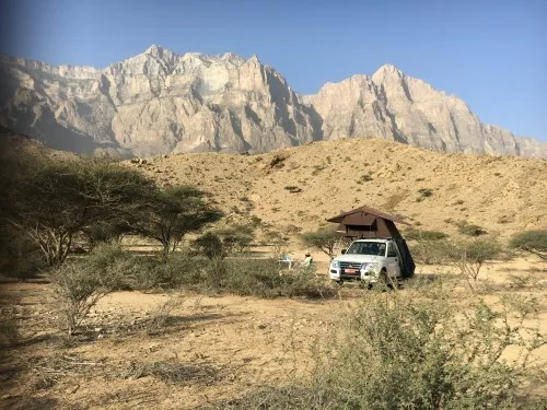 A white camper truck with a pop-up roof is parked on a dry, rocky plain with sparse bushes. A person sits on a chair nearby, with tall, rugged mountains dominating the background under a clear blue sky.