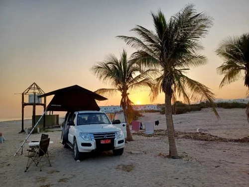 A white SUV with a rooftop tent is parked on a sandy beach near palm trees at sunset. Camping chairs and a table are set up nearby, and buildings are visible in the background.
