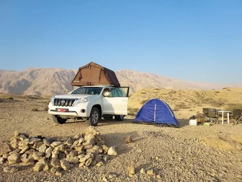 A white SUV with a rooftop tent is parked on rocky desert terrain beside a blue ground tent. Camping gear and a table are set up nearby, with mountains in the background under a clear blue sky.