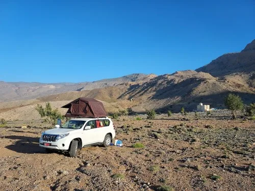A white SUV with a rooftop tent is parked on rocky terrain under a clear blue sky, surrounded by mountains and sparse vegetation. Camping supplies are on the ground next to the vehicle.
