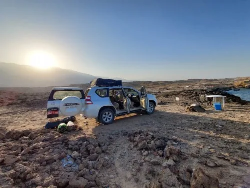 A white SUV with open doors is parked on rocky terrain at sunrise or sunset. Camping gear is spread out nearby, and a small table with supplies is set up by the edge of the rocky landscape. Distant mountains are visible.