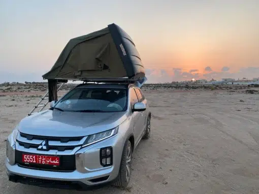 josh-oman-4x4-rental (1) A silver Mitsubishi SUV with an Omani license plate is parked on a sandy landscape at sunset. A tent is set up on the roof of the car, with a ladder for access. The sky is partly cloudy with orange hues.
