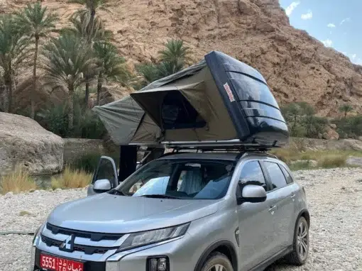 josh-oman-4x4-rental (14) A silver Mitsubishi SUV with a red license plate is parked on rocky ground near cliffs and palm trees. A tent is open on the roof of the car, set up for camping under a partly cloudy sky.