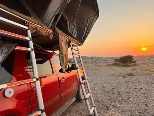 josh-oman-4x4-rental (15) A red SUV with tents and ladders on the roof is parked on a rocky field at sunset. The sky is orange and clear, and the sun is low on the horizon, casting a warm light over the landscape.