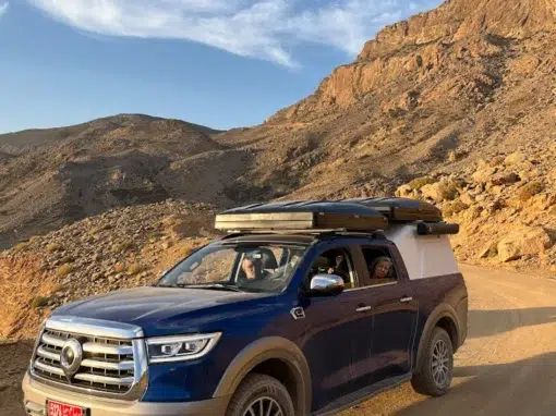 josh-oman-4x4-rental (16) A blue van with a covered loading area is parked on a dirt road in a rocky, mountainous landscape, under a clear sky. The driver is visible inside the vehicle.