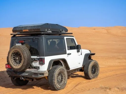 A white off-road SUV, equipped with a roof-mounted storage box and rear-mounted spare wheel, is parked on sandy desert terrain under a clear blue sky.