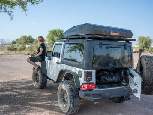 A person sits on the front fender of a parked Jeep, with the rear door and roof box open, in a dry, open landscape, with scattered trees and distant mountains under a clear sky.