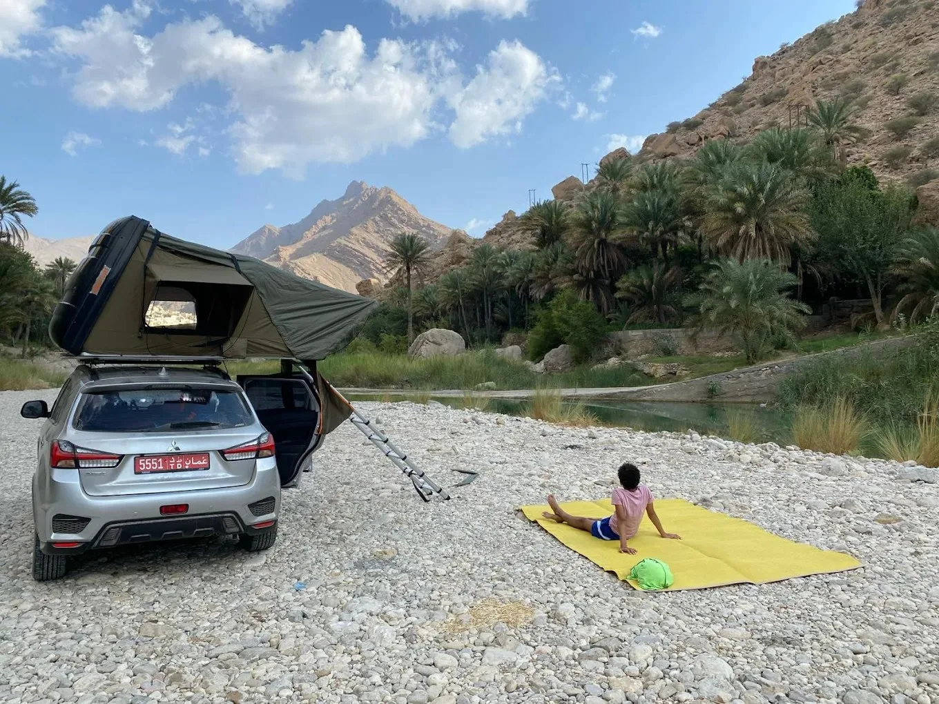 A car with a tent on the roof is parked on rocky ground near a river, surrounded by palm trees and mountains. A child sits on a yellow carpet next to the car, under a partly cloudy sky.