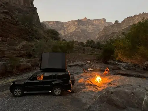 josh-oman-4x4-rental (7) A black SUV with a tent on the roof is parked on rocky ground near a small campfire, surrounded by cliffs and mountains at sunset. Two people are sitting by the fire under a clear sky.