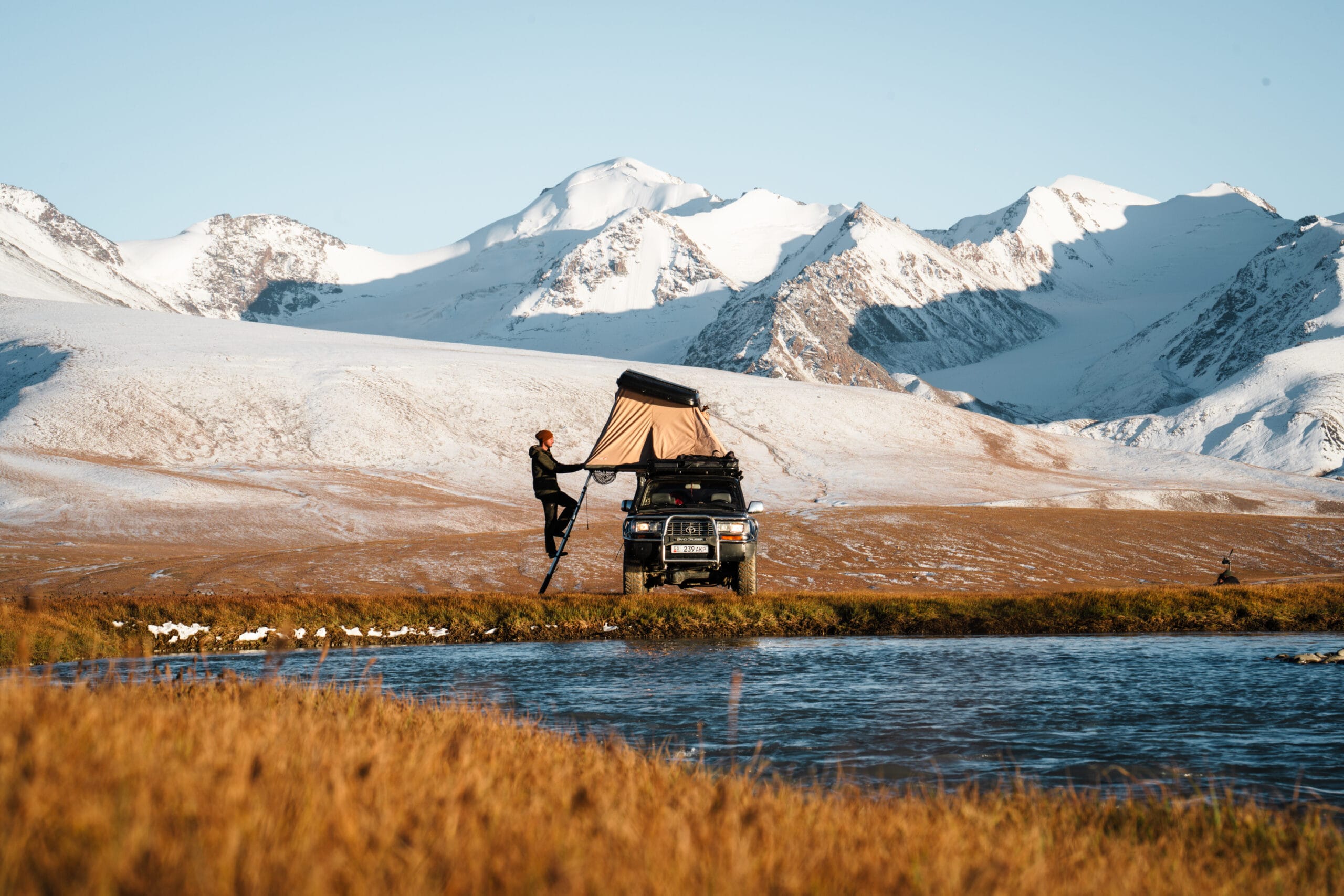 Une personne se tient sur une échelle à côté d'un véhicule utilitaire sport avec une tente sur le toit, installé près d'un ruisseau dans un champ herbeux. Des montagnes enneigées s'élèvent à l'arrière-plan sous un ciel bleu clair.