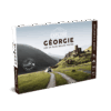 Le Guide de voyage Explore Géorgie présente une image panoramique d'une voiture se dirigeant vers d'anciennes ruines en pierre dans des collines verdoyantes, avec des montagnes majestueuses à l'arrière-plan.
