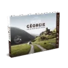 Le Guide de voyage Explore Géorgie présente une image panoramique d'une voiture se dirigeant vers d'anciennes ruines en pierre dans des collines verdoyantes, avec des montagnes majestueuses à l'arrière-plan.