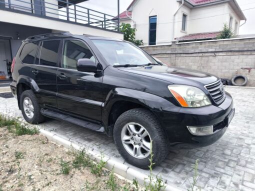 vlad-4x4-rental-bishkek-lexus (11) A black SUV with silver rims is parked on a paved driveway next to a stone wall, with a modern house visible in the background.