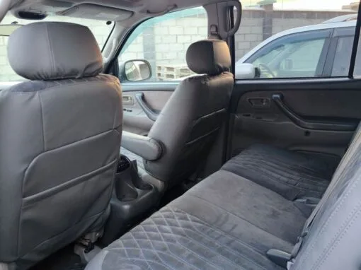 The image shows the interior of a car, focusing on the rear seat with gray upholstery. The front seats are visible and the car door is opened, revealing part of a brick wall and another vehicle outside.