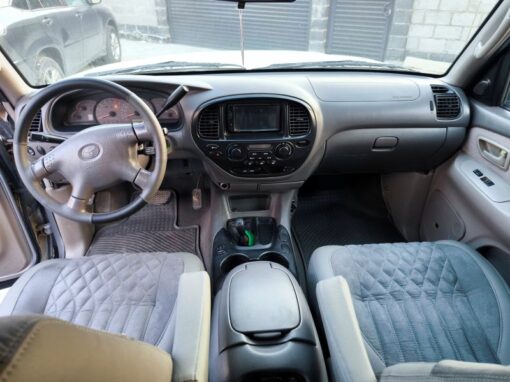 vlad-4x4-rental-bishkek-lexus (3) The interior of a car showing the dashboard, steering wheel, center console and two front seats with padded covers. Sunlight streams in through the windscreen and a building is visible outside.