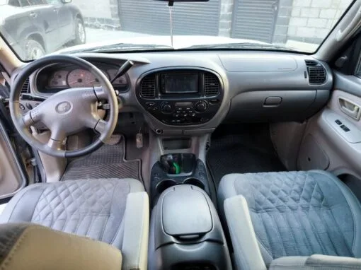 The interior of a car showing the dashboard, steering wheel, center console and two front seats with padded covers. Sunlight streams in through the windscreen and a building is visible outside.