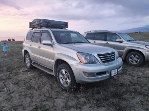 vlad-4x4-rental-kyrgyzstan-Lexus GX470 (1) A silver Lexus SUV with a roof-mounted cargo box is parked in a grassy area next to another SUV. A child stands nearby, with a body of water and a cloudy sky in the background at sunset.