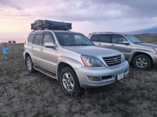 A silver Lexus SUV with a roof-mounted cargo box is parked in a grassy area next to another SUV. A child stands nearby, with a body of water and a cloudy sky in the background at sunset.