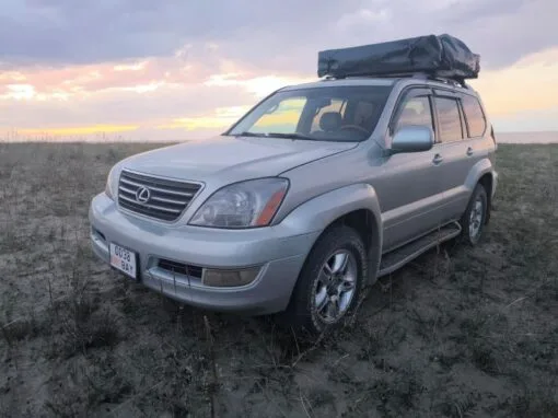 A silver Lexus SUV with a roof box is parked on dry grass with a cloudy sky and sunset in the background.