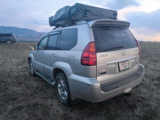 vlad-4x4-rental-kyrgyzstan-Lexus GX470 (7) A silver Lexus SUV with a roof rack is parked on a grassy field, with mountains and another vehicle visible in the background under a cloudy sky.