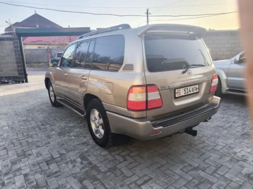 A silver Toyota Land Cruiser V8 SUV is parked on a paved driveway next to a brick wall. Another vehicle is partially visible to the right.
