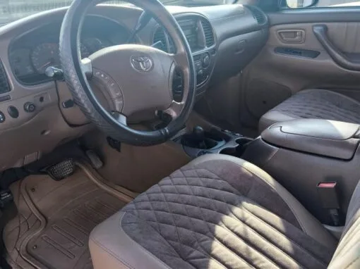The interior of a Toyota vehicle showing the steering wheel, dashboard, gearshift and front seats with padded covers. Sunlight streams through the windows, and a concrete wall is visible on the outside.