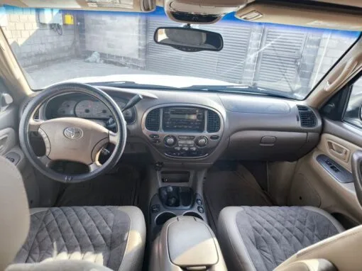 Interior view of a Toyota SUV, showing the steering wheel, dashboard, center console and front seats with padded covers. Sunlight fills the cabin, and the vehicle is parked next to a gray brick wall outside.