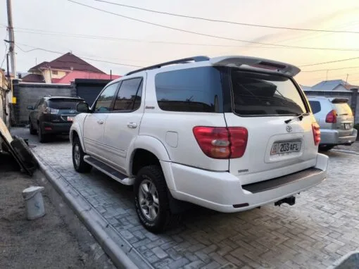 A white Toyota SUV is parked on a paved driveway near a house, with other vehicles and buildings visible in the background as the sun sets.