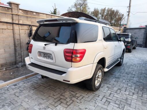 vlad-4x4-rental-kyrgyzstan-sequoia (11) A white Toyota SUV is parked on a paved driveway next to a concrete wall, with another car visible in the background. The area appears to be residential, with a few trees and power poles nearby.