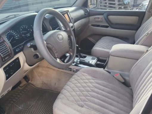 The interior of a Toyota Land Cruiser with gray quilted fabric seats, gray steering wheel, center console controls and sleek dashboard. The sun shines through the windows and a wall of pallets is visible outside.