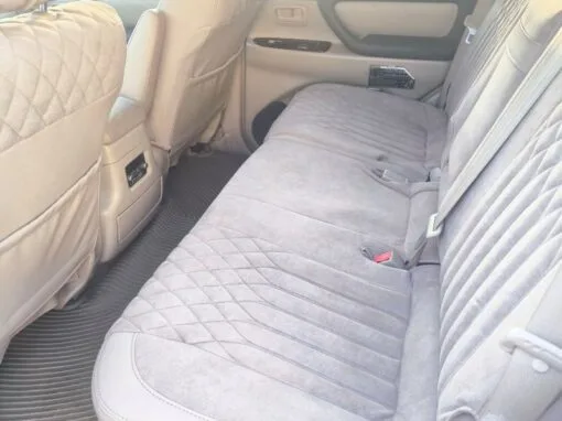 The image shows the back seat of a car with light gray quilted upholstery, floor mats and sunlight streaming in from the left side. The car door is closed and the windows reveal an exterior brick wall.