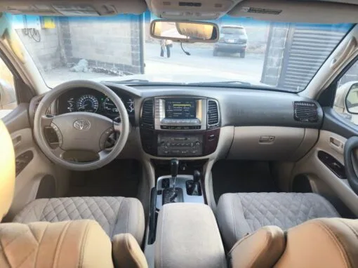 Interior view of a Toyota vehicle, showing the steering wheel, dashboard with illuminated gauges, central infotainment system, gearshift lever and padded beige seats. A partially open gate and cars are visible outside the windscreen.