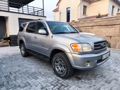 A silver SUV is parked on a paved driveway next to a building with beige walls and a fence. Several stacked tires are visible in the background. The front and side of the car are clearly represented.