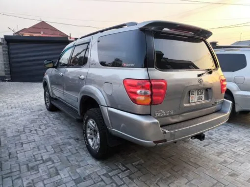 A silver Toyota Sequoia SUV is parked on a paved surface near a black gate and a red-roofed building, with another vehicle visible on the right-hand side.