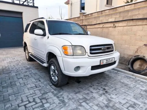 A white Toyota Sequoia SUV is parked in a paved driveway next to a brick wall and garage door, with buildings visible in the background. The vehicle's license plate reads 203 AFL.